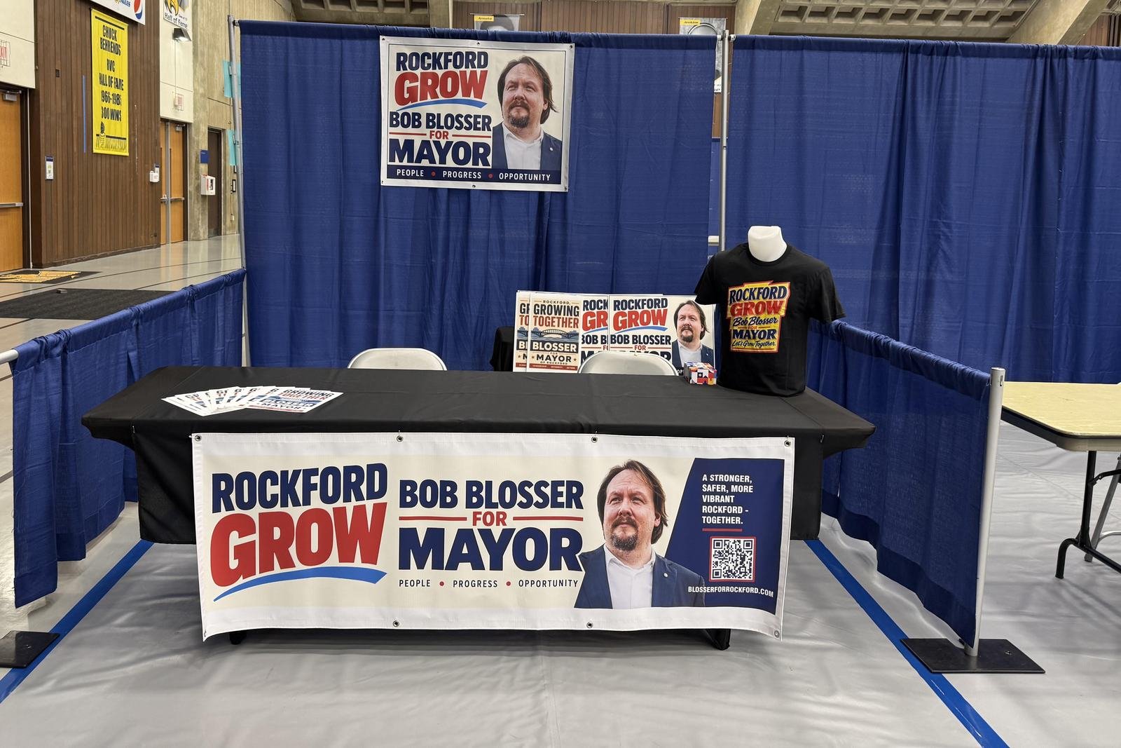 The campaign&rsquo;s exhibitor table set up on the gym floor at Rock Valley College: a black-draped table with a &ldquo;Rockford Grow &mdash; Bob Blosser for Mayor&rdquo; banner across the front, a matching banner hung on the blue curtain behind it, literature fanned out on the table, and a campaign T-shirt on a display form at one end.