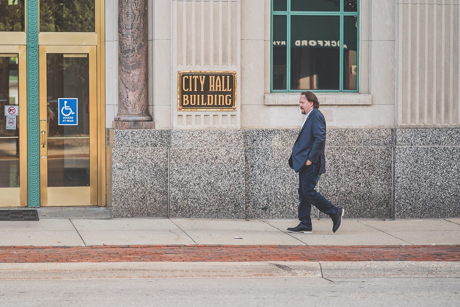 Bob Blosser walking along the sidewalk past the brass City Hall Building sign on the granite front of Rockford City Hall.