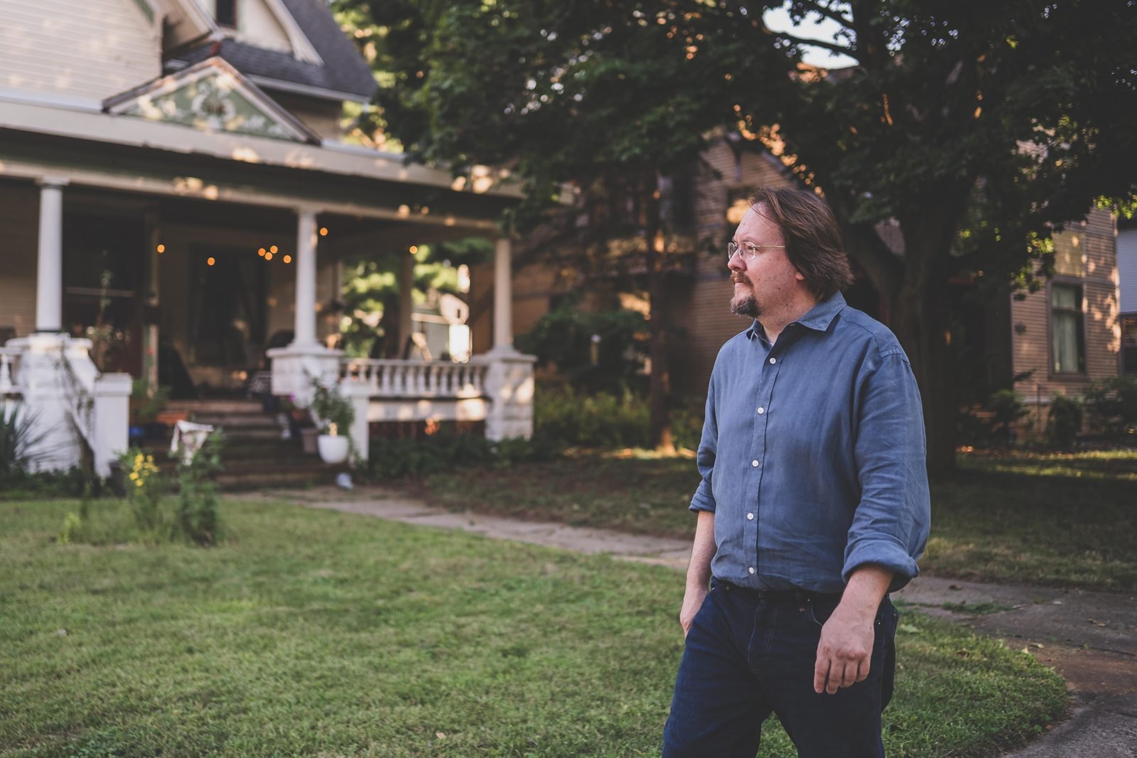 Bob Blosser walking across a front lawn at dusk, past the porch of an older Rockford house strung with lights.