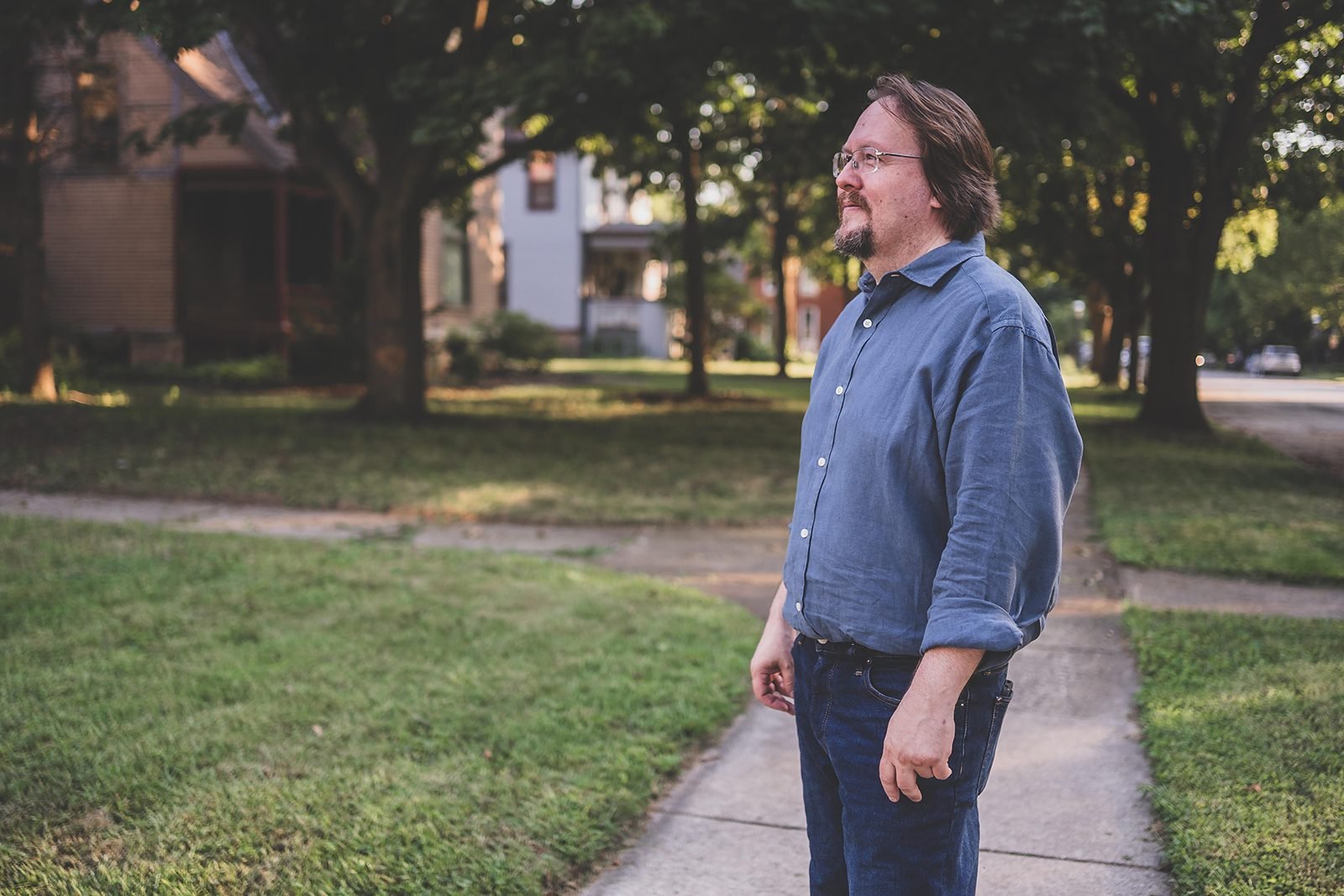 Bob Blosser standing on a residential sidewalk in Rockford, with houses, front porches and mature trees along the block behind him.