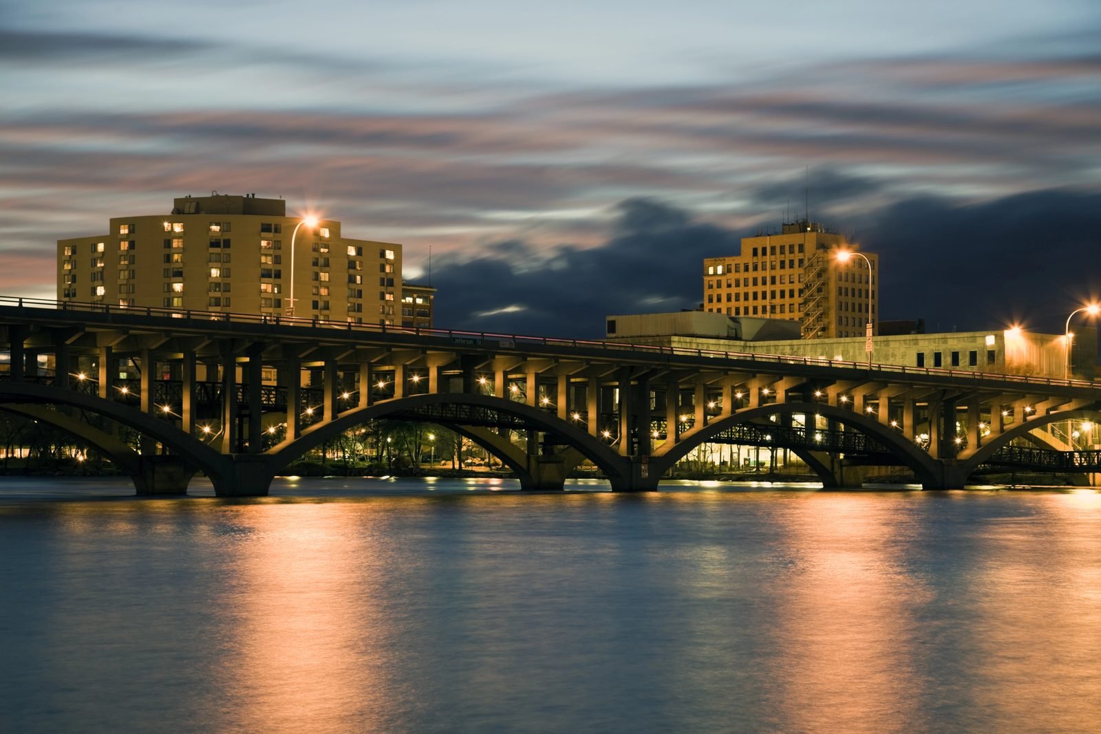 The concrete arches of the Jefferson Street bridge seen from water level at dusk, receding toward the lit downtown Rockford bank.