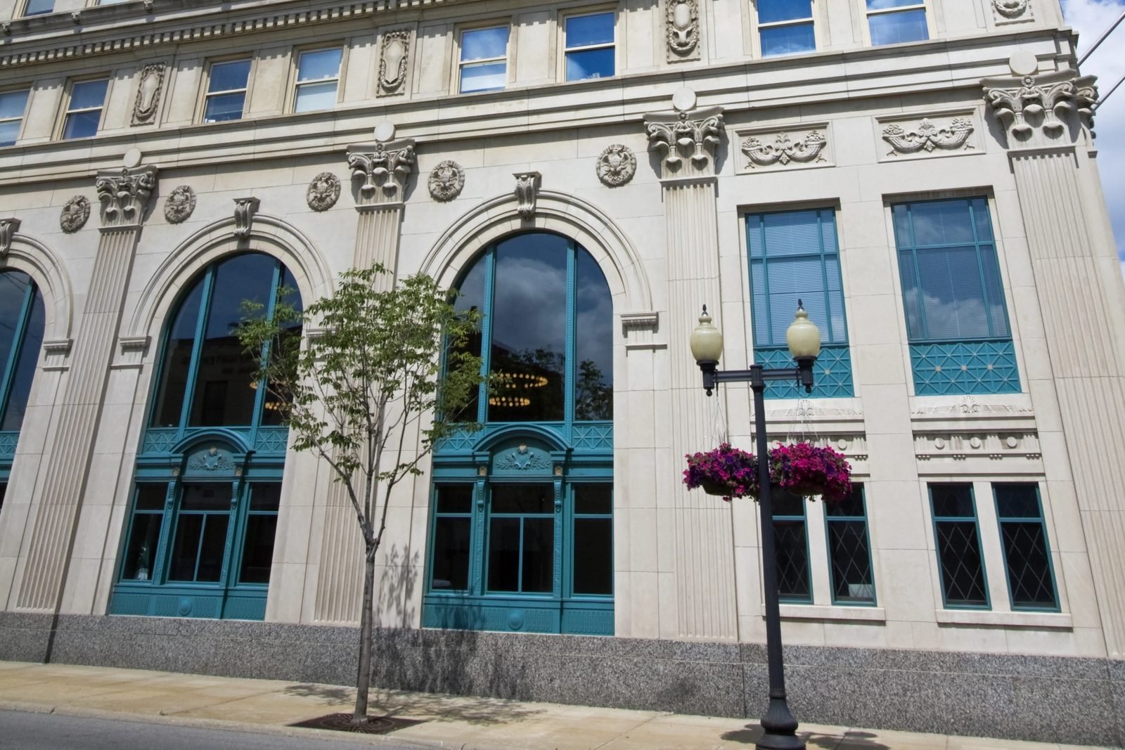 The street-level facade of Rockford City Hall: limestone arches with teal-green joinery, a granite base, a young tree and a lamppost hung with flowers.