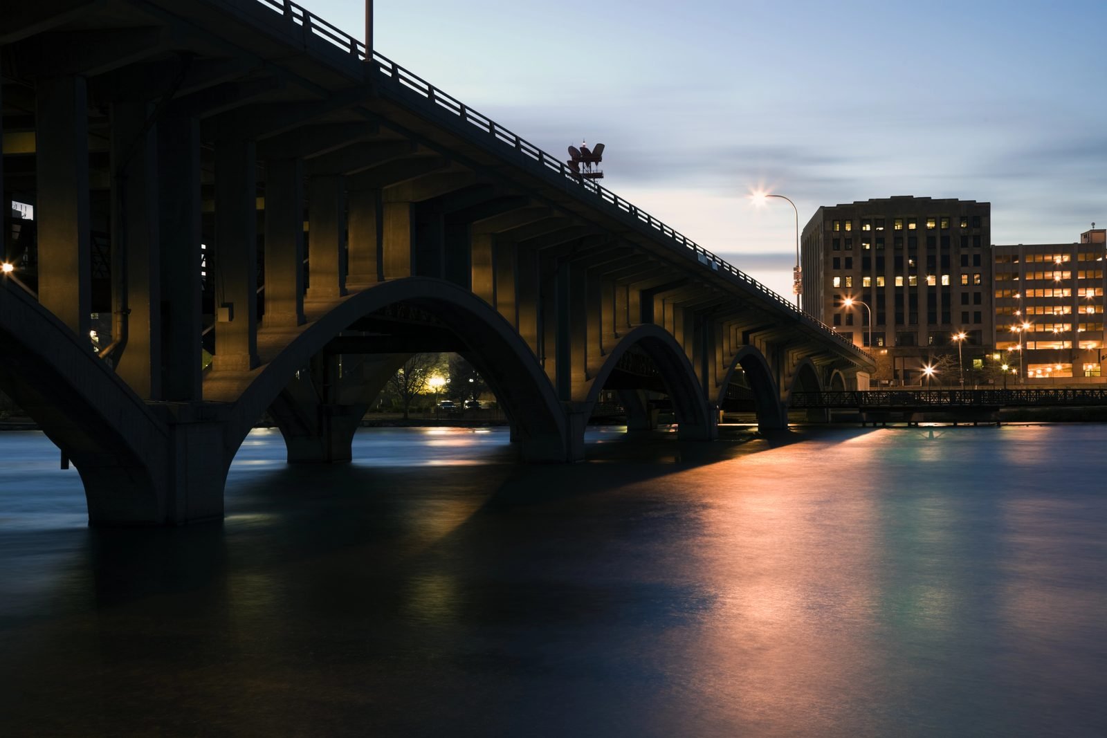 The Jefferson Street bridge over the Rock River at dusk, lit along its length, with the downtown Rockford skyline behind it.