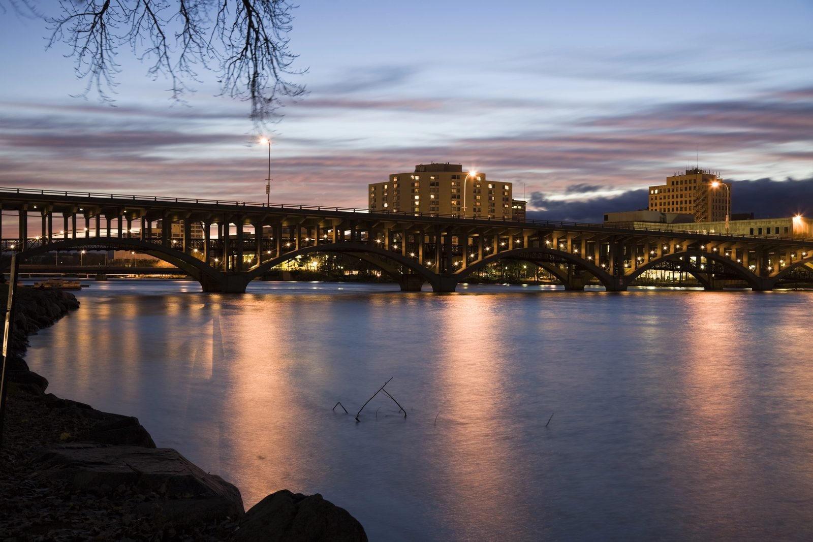 The Jefferson Street bridge over the Rock River at dusk, its arches and lamps reflected in the water, downtown Rockford lit behind it.