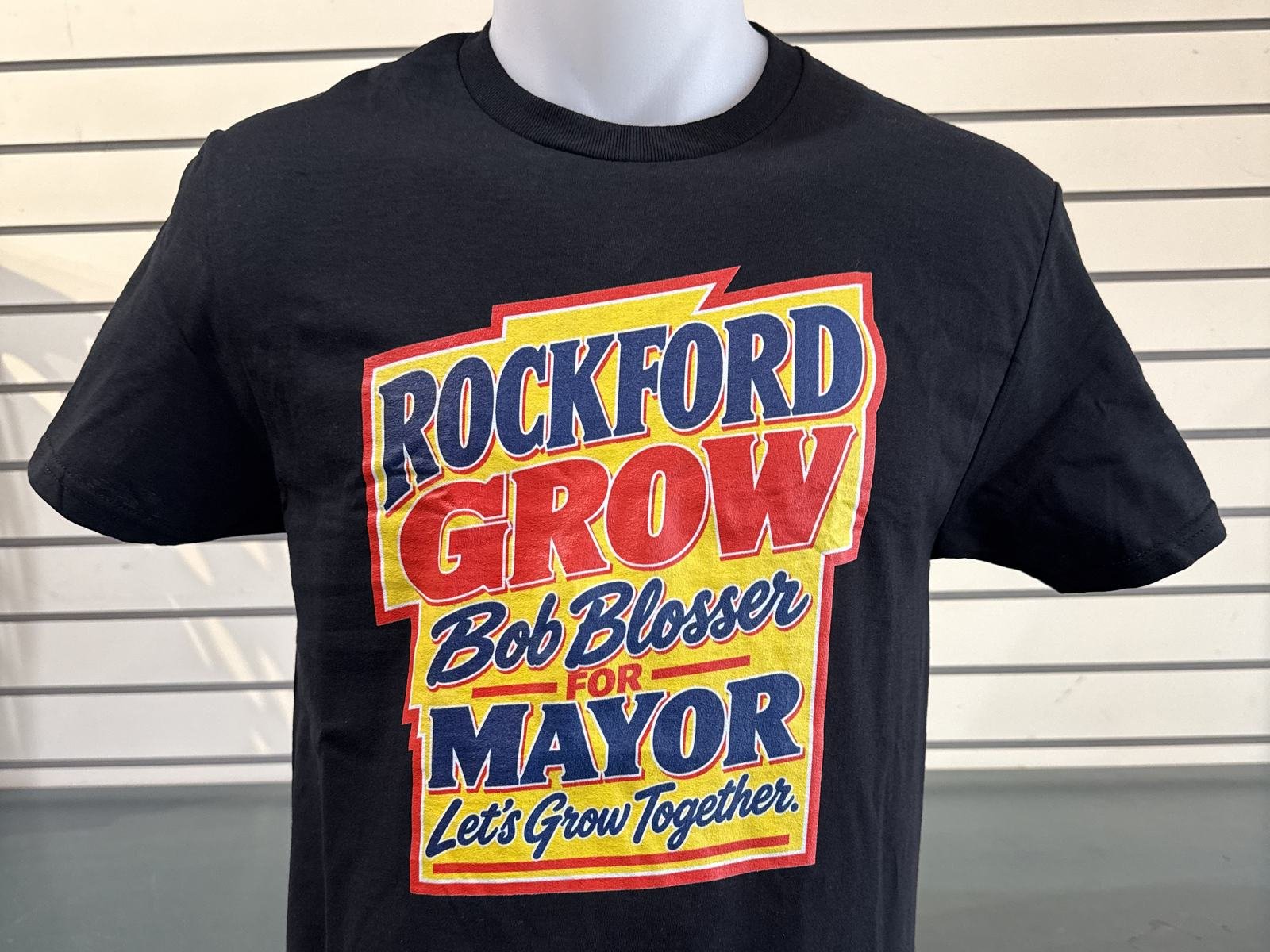 A black T-shirt on a display form, printed with a yellow sign-painter panel outlined in red reading &ldquo;Rockford Grow, Bob Blosser for Mayor, Let&rsquo;s Grow Together&rdquo; in navy and red lettering.
